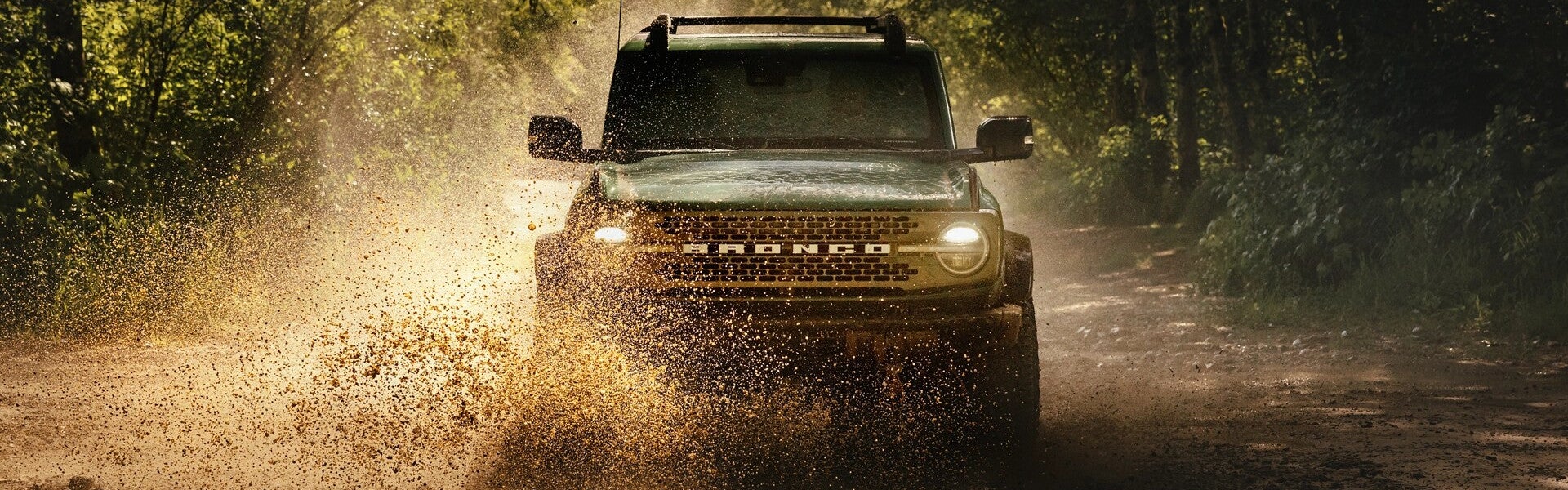 An off-road Ford Bronco splashes through muddy forest trail with water and dirt flying all around.