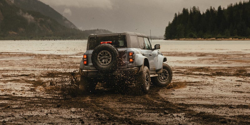 An off-road Ford Bronco splashes through muddy forest trail with dirt flying all around.